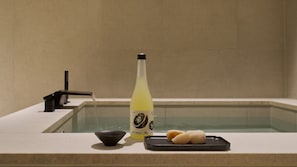 Una botella de sake, un tazón pequeño y una bandeja con copas de sake sobre una encimera junto a una bañera.