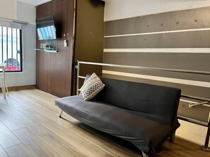 Een kamer met een grijze bank, een houten kledingkast, een televisie aan de muur en een raam met uitzicht op een speeltuin.