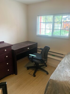 Ein Zimmer mit Schreibtisch, Stuhl, Kommode und einem Fenster mit Jalousien.