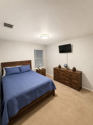 Ein Schlafzimmer mit Bett, Kommode, Fernseher und einem Fenster mit Jalousien.