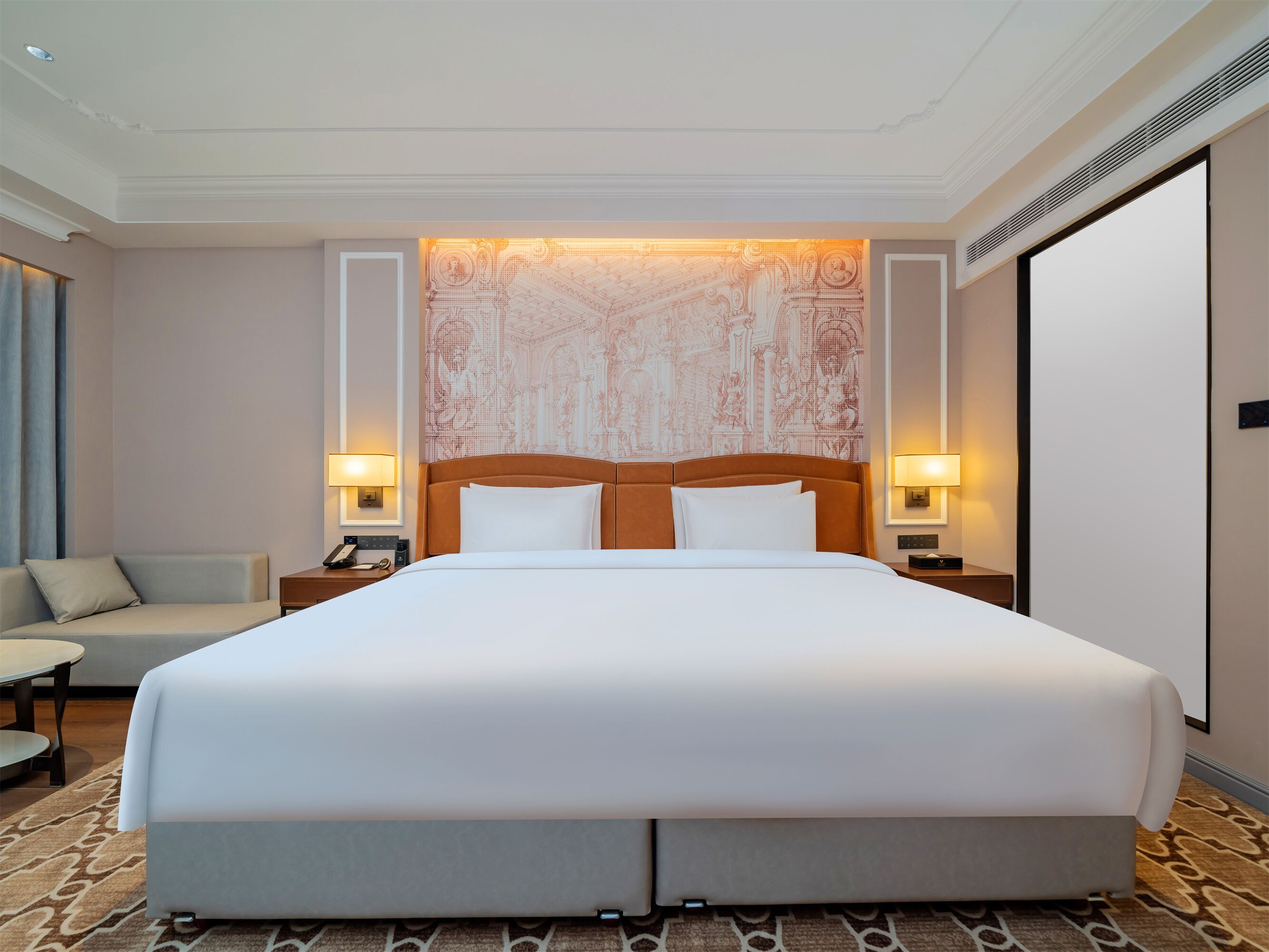 Una habitación de hotel con una cama grande, dos mesitas de noche con lámparas, un sofá y una alfombra estampada.