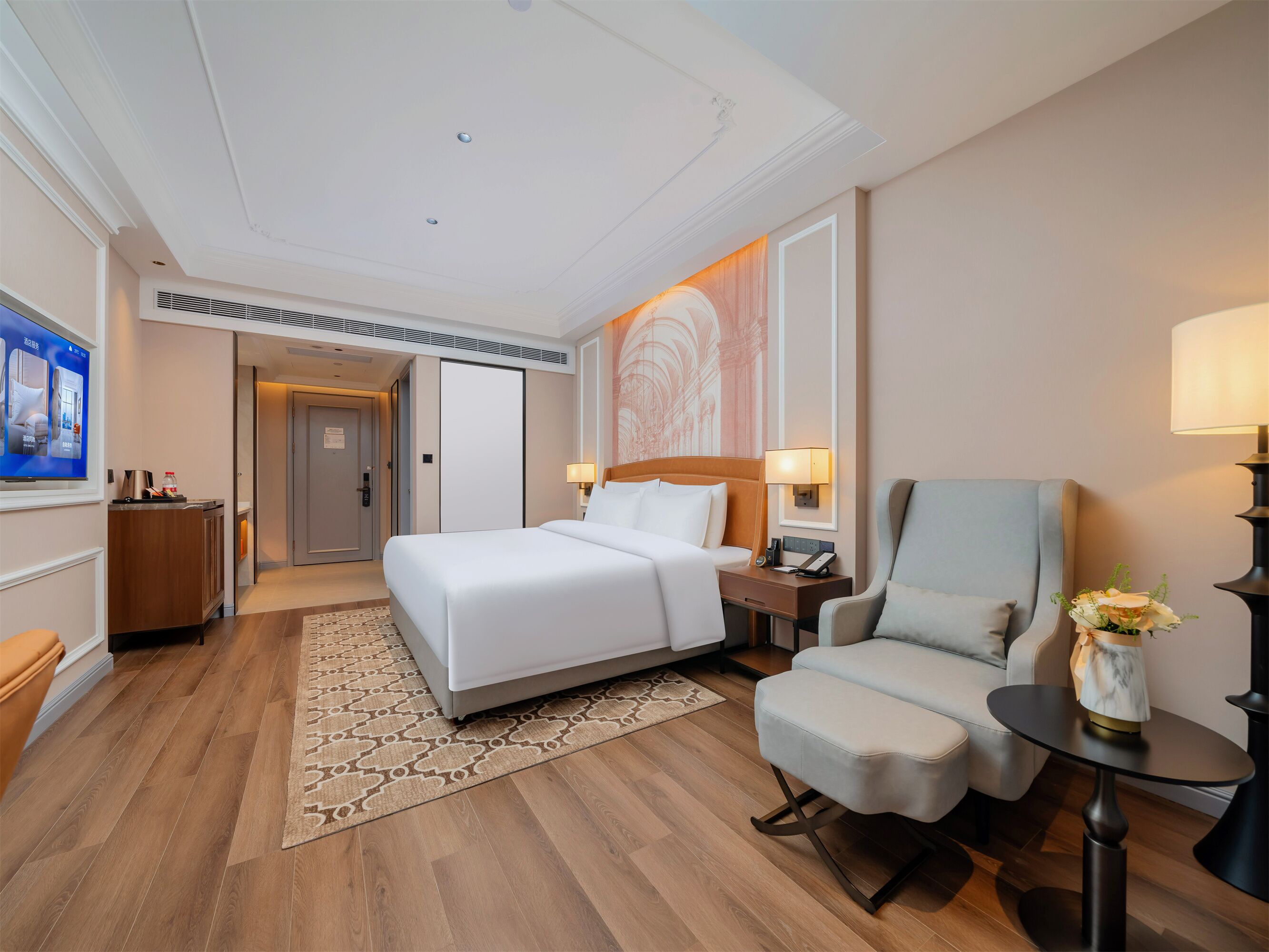 Habitación de hotel con cama, una silla, escritorio, lámpara, un jarrón con flores, televisor y puerta.