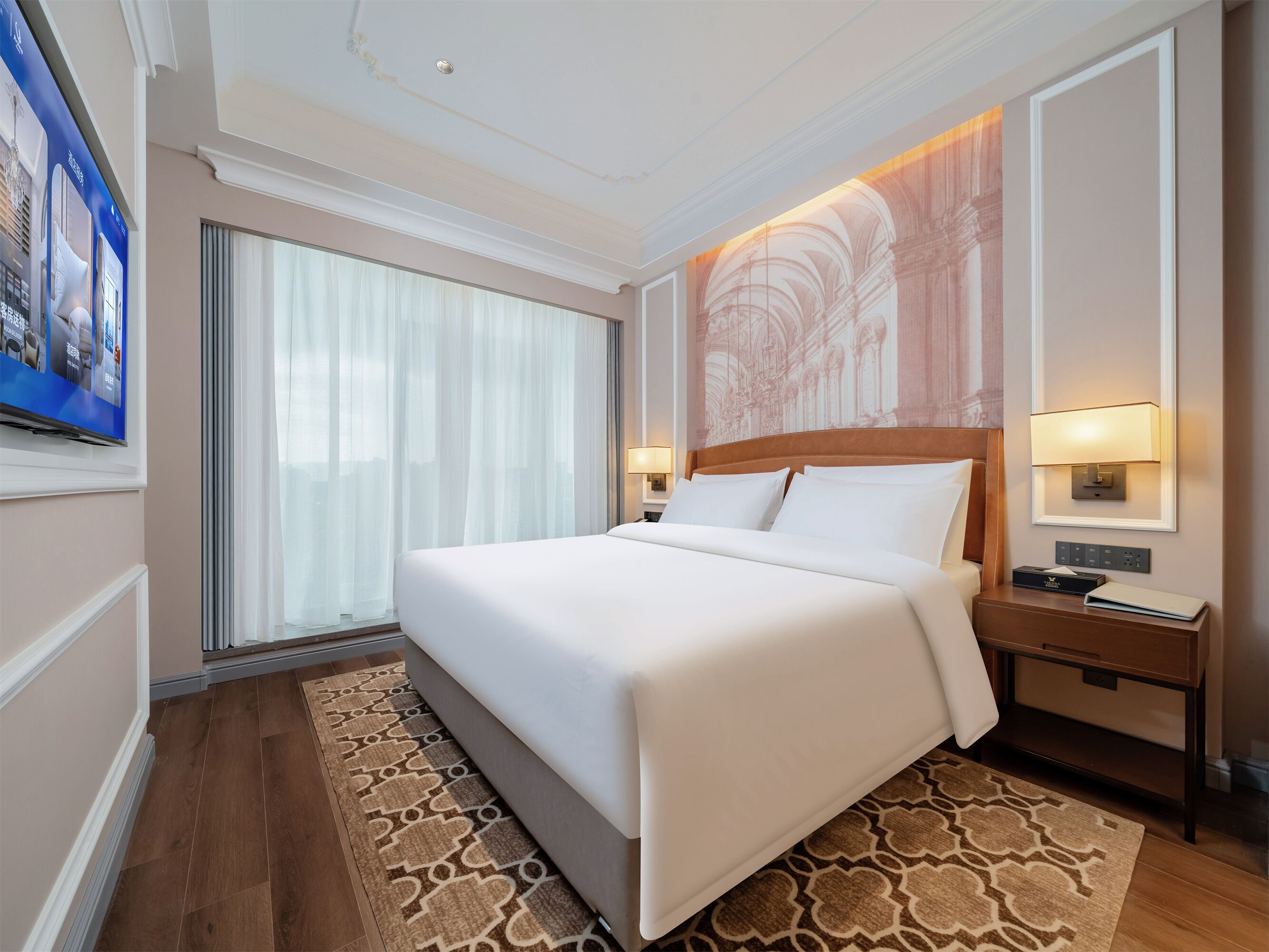 Quarto de hotel com uma cama grande, mesa de cabeceira, luminária fixada na parede e uma janela ampla com cortinas leves.