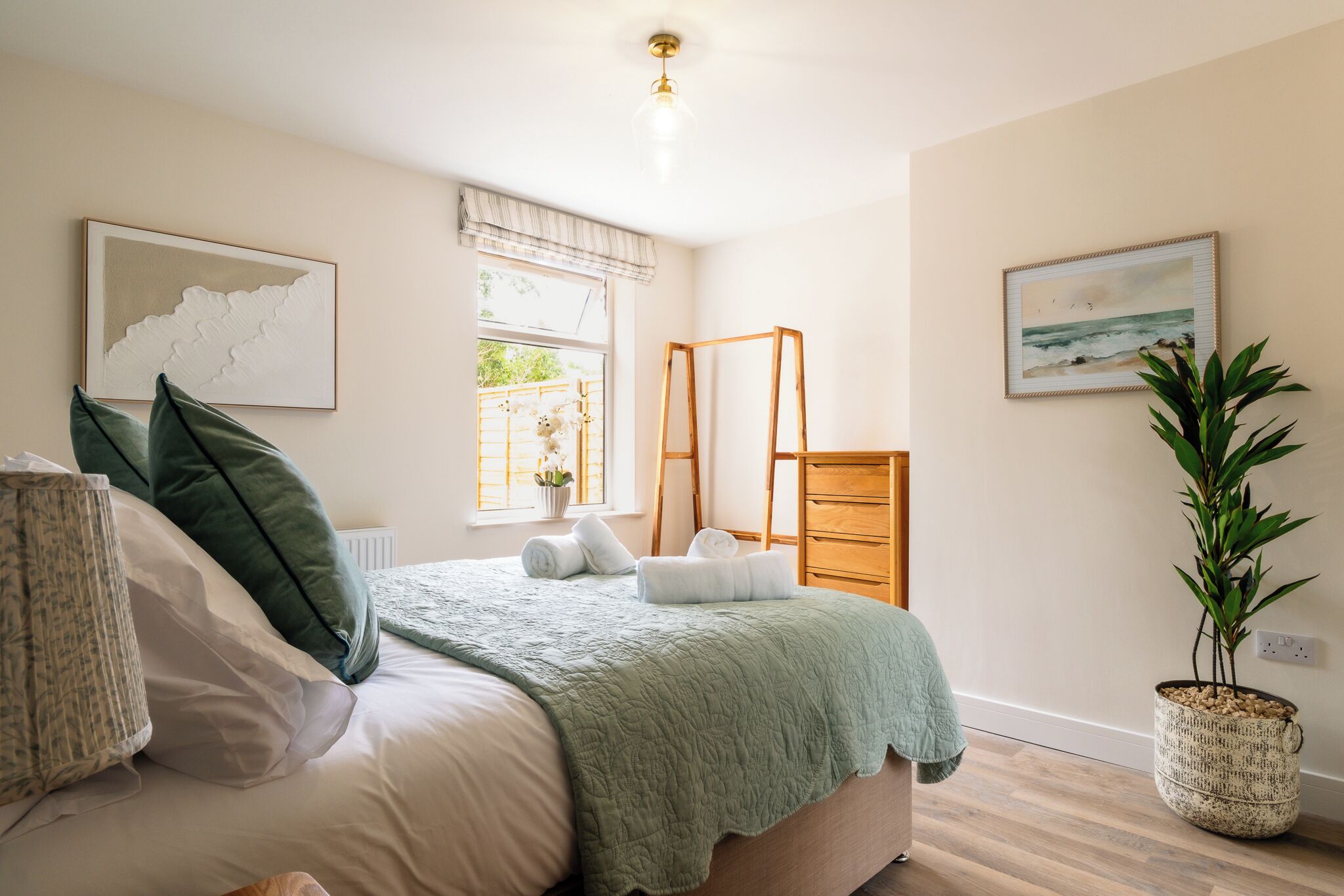 A bedroom with a bed, pillows, a green blanket, a window with a view, a ladder, a plant, and artwork on the walls.