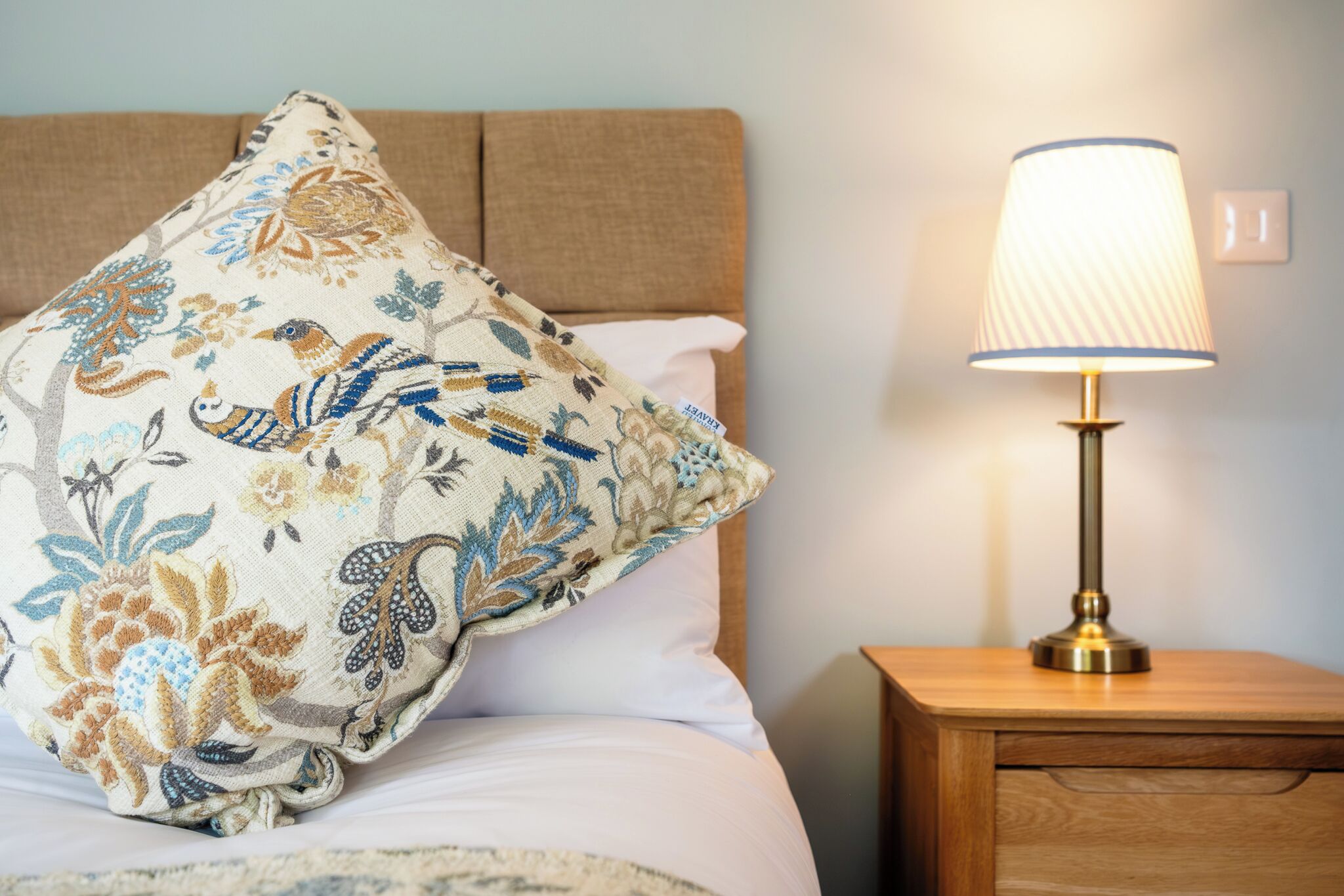 Una cama con un cojín floral, una lámpara de noche y una mesita de noche de madera.