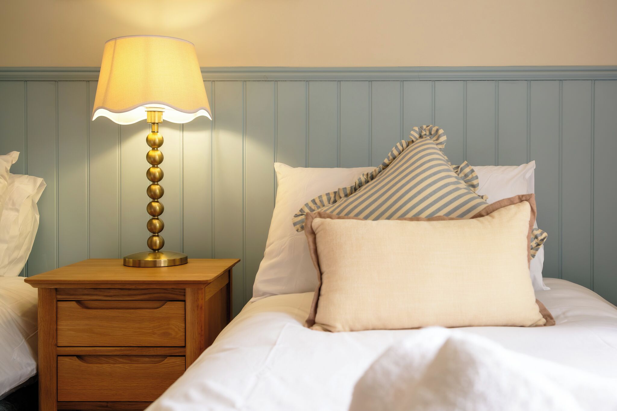 A bedroom with a wooden nightstand, a lamp, a bed with striped pillows, and a blue paneled headboard.