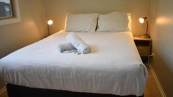 Una cama bien hecha con sábanas blancas, dos lámparas de noche y una mesita auxiliar pequeña.
