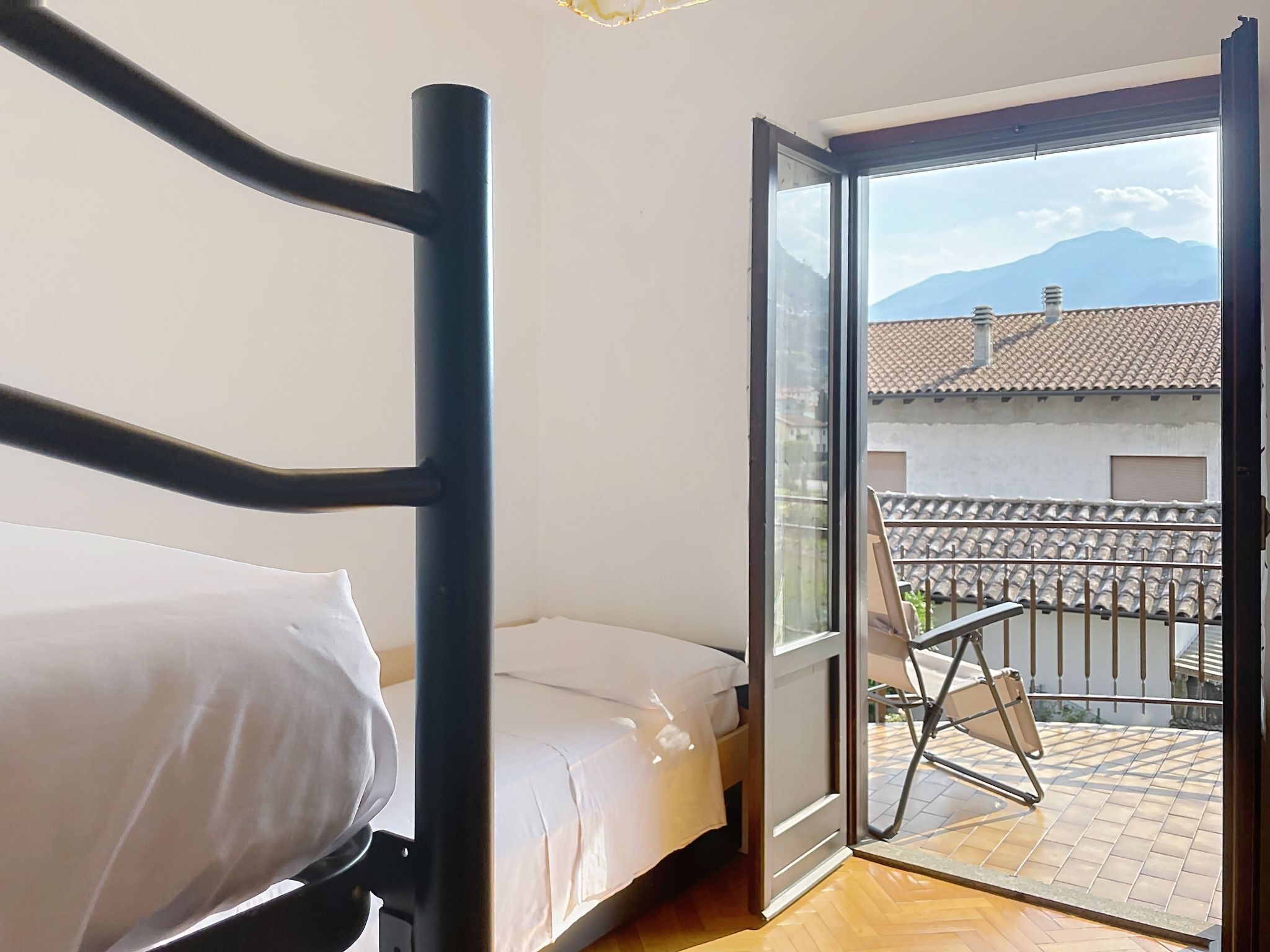 A room with a bed, a chair, and a balcony with a view of buildings and mountains.