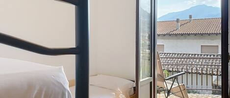 A room with a bed, a chair, and a balcony with a view of buildings and mountains.