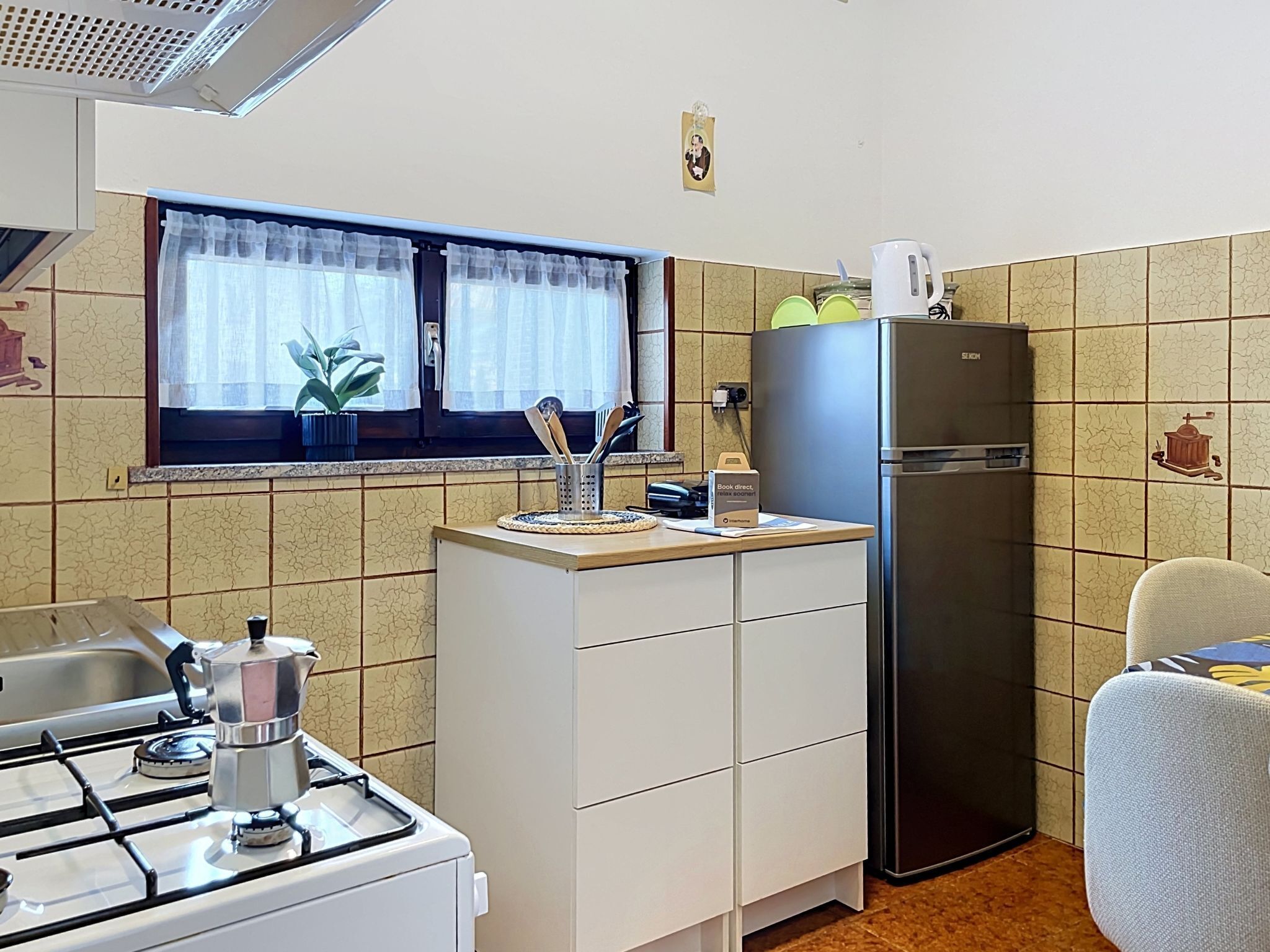 Eine kleine Küche mit Gaskochfeld, Kühlschrank und einem Fenster mit Vorhängen.