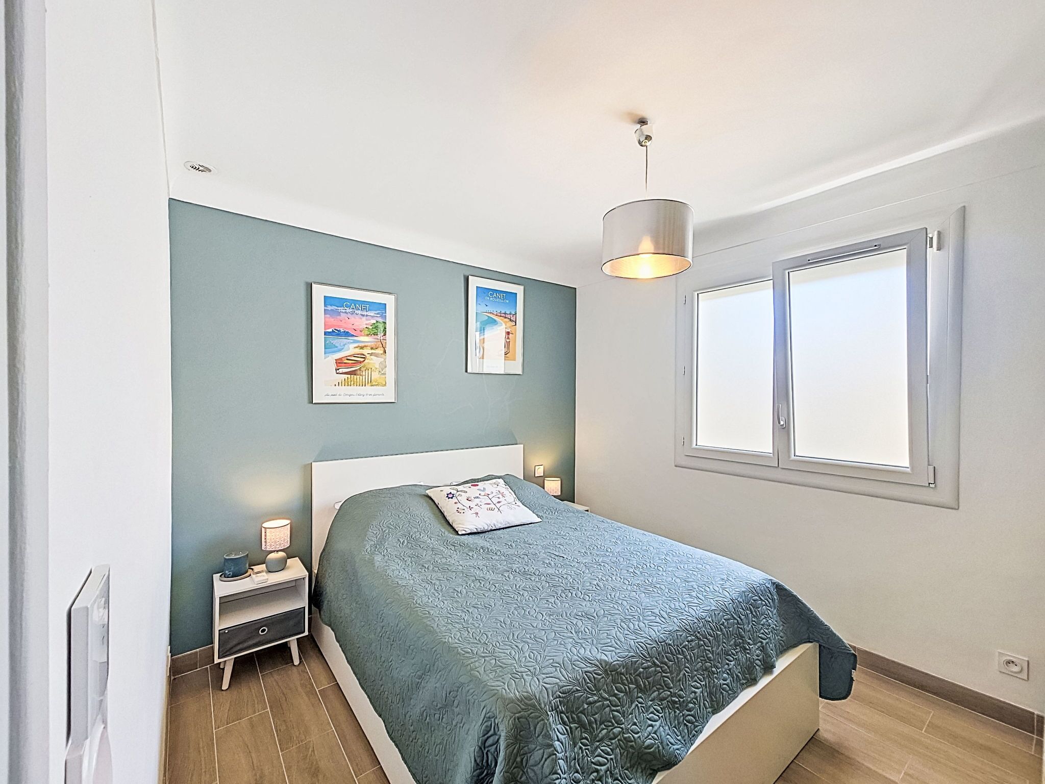 A bedroom with a bed, a nightstand, a lamp, two framed pictures, a window, and a hanging light fixture.