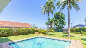 A swimming pool with a net, surrounded by a paved patio, palm trees, and a hedge. - Romsey Holiday Breeze (Durban North)