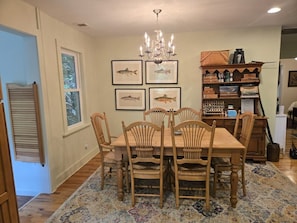 Comedor con mesa y sillas de madera, candelabro, ilustraciones enmarcadas de peces y una estantería con diversos objetos.