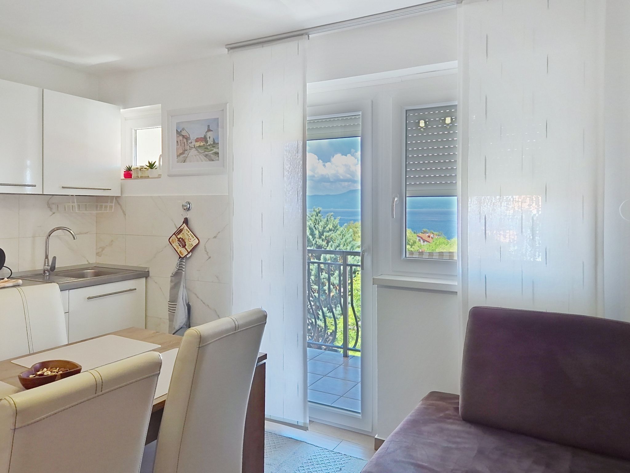 A modern kitchen with white cabinets, a dining area with a table and chairs, and a balcony with a view of the sea.