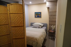Een slaapkamer met een bed, een nachtkastje met een vaas, een foto aan de muur en een geweven scheidingswand.