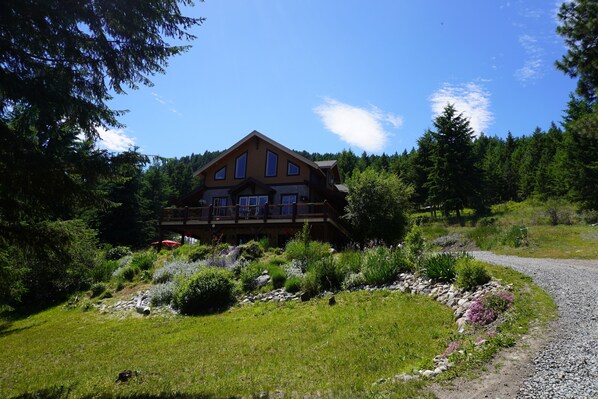 A large wooden house with multiple windows and a balcony, surrounded by lush greenery and a gravel path.