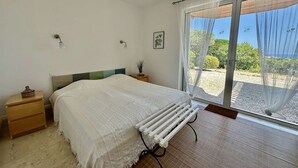 Ein Schlafzimmer mit Bett, Heizkörper, Nachttisch, Wandlampe und einem Fenster mit Blick ins Grüne.