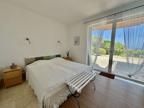 Ein Schlafzimmer mit Bett, Heizkörper, Nachttisch, Wandlampe und einem Fenster mit Blick ins Grüne.