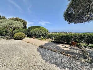 Ein Schotterweg führt zu einem Haus mit Meerblick und einem gepflegten Garten mit verschiedenen Sträuchern und Bäumen.