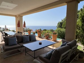 Eine überdachte Terrasse mit Tisch und Sitzgelegenheiten mit Blick auf einen Pool und das Meer.