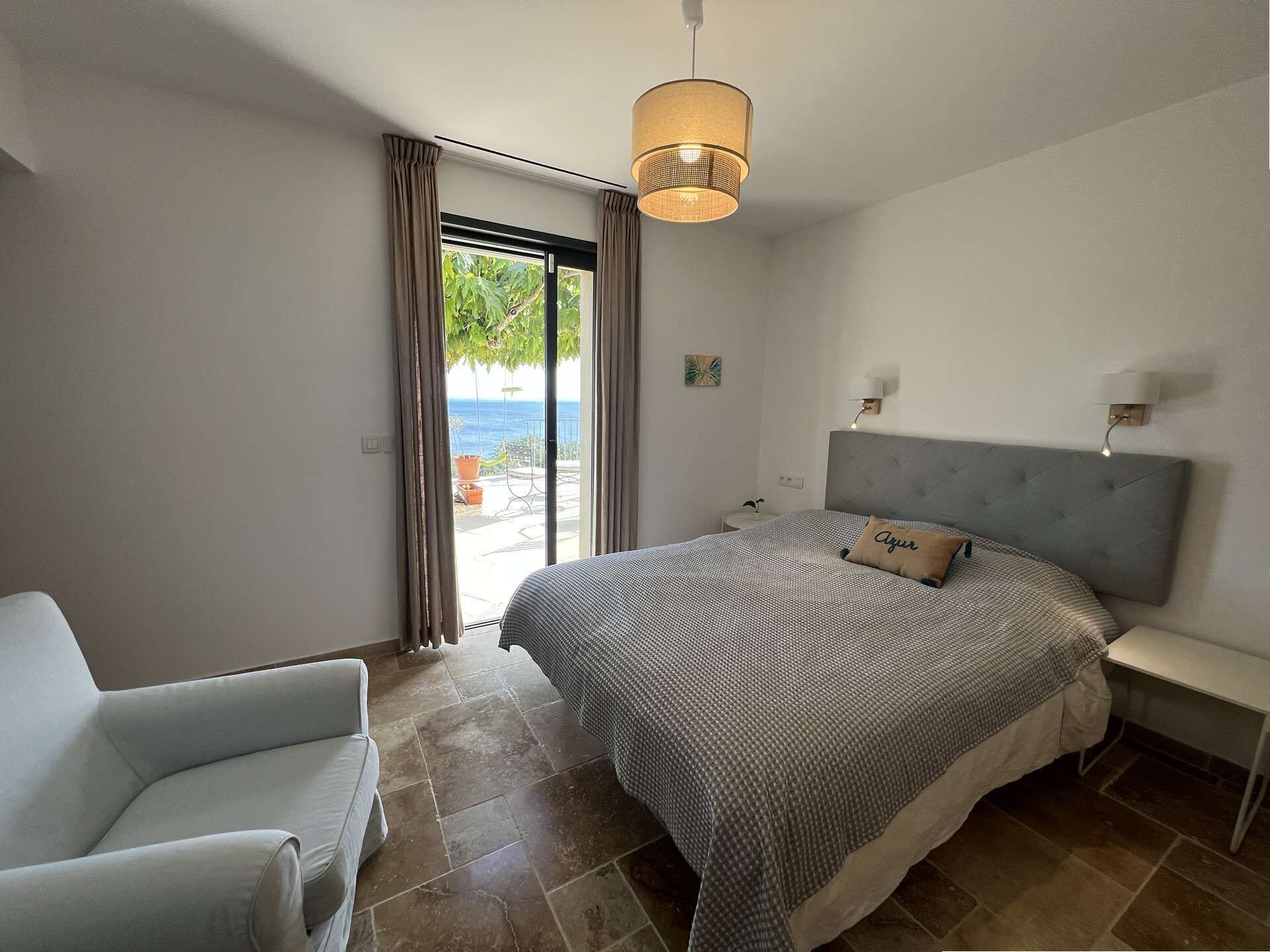 Une chambre à coucher comprenant un lit, une chaise, une table de chevet, une lampe et une vue sur la mer, accessible par une porte coulissante.