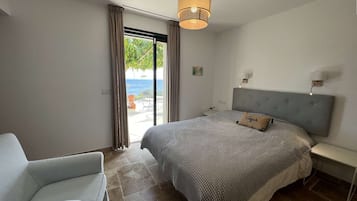 Quarto com cama, cadeira, mesa de cabeceira, abajur e vista para o mar por uma porta de correr.