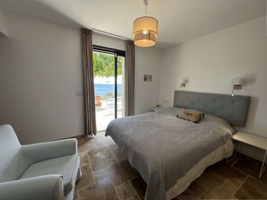 Ein Schlafzimmer mit Bett, einem Sessel, einem Nachttisch, einer Lampe und Meerblick durch eine Schiebetür.