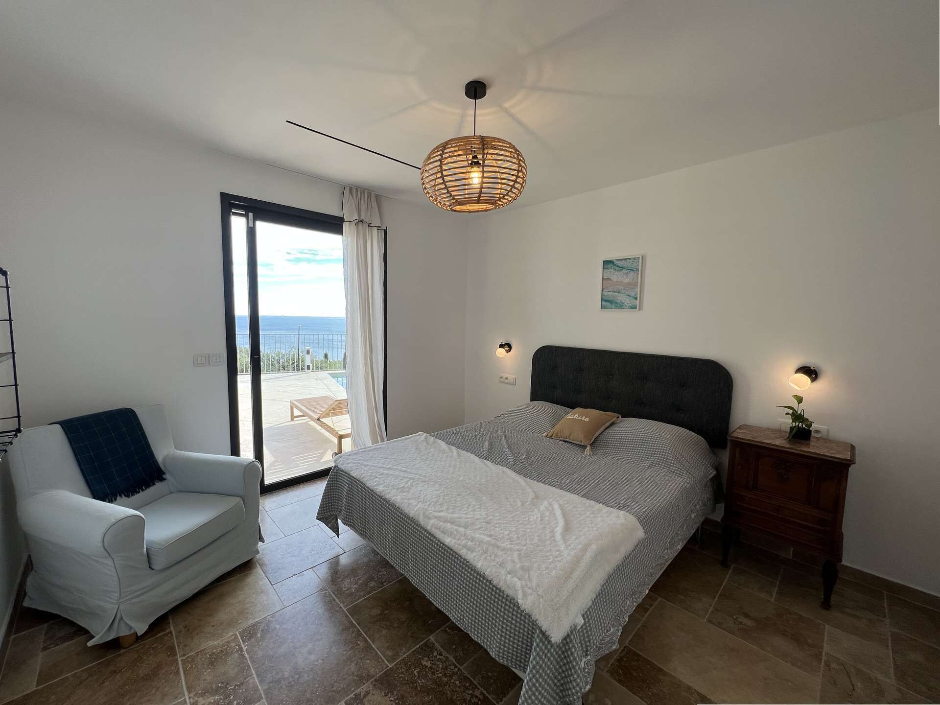 Un dormitorio con cama, una silla, una mesita de noche, una lámpara de pared, un ventilador de techo y vistas al mar a través de una puerta corrediza.