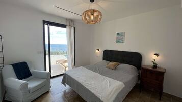 Un dormitorio con cama, una silla, una mesita de noche, una lámpara de pared, un ventilador de techo y vistas al mar a través de una puerta corrediza.