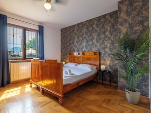 Uma cama de madeira com cabeceira, uma janela com cortinas, um radiador, uma pequena mesa com abajur e um vaso de planta.