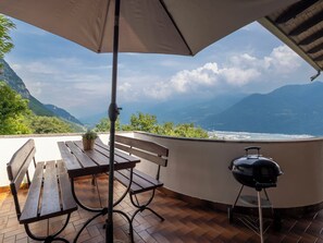 Eine Terrasse mit Holztisch und Bank, einem Grill und Blick auf eine gebirgige Küstenlandschaft.