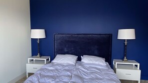 Ein Schlafzimmer mit einem dunkelblauen Kopfteil, weißer Bettwäsche und zwei Nachttischen mit Lampen.