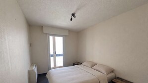A bedroom with a bed, a nightstand, a small table, a ceiling light, and a sliding door to a balcony. - SAINT-RAPHAËL CITY CENTER – BEACH WITHIN WALKING DISTANCE – COMFORTABLE 1-BEDROOM APARTMENT (ST RAPHAEL)