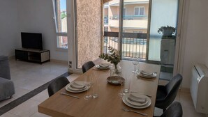 A dining area with a wooden table set for four, a TV in the corner, and a view of a balcony with a railing. - SAINT-RAPHAËL CITY CENTER – BEACH WITHIN WALKING DISTANCE – COMFORTABLE 1-BEDROOM APARTMENT (ST RAPHAEL)