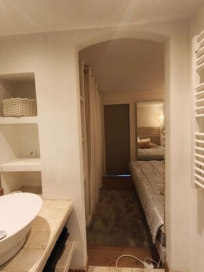 Ein schmaler Flur führt zu einem Schlafzimmer mit Bett, einer kleinen Ablage mit Korb und einem Badezimmer mit Waschbecken.