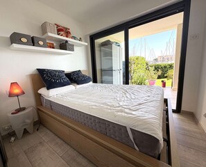 Ein Schlafzimmer mit Bett, Nachttischlampe, kleinem Tisch und Blick auf Grünflächen und Gebäude.