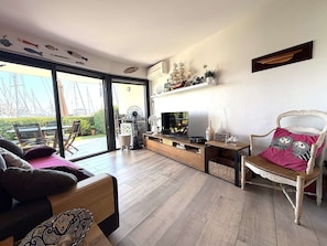 Ein geräumiger Wohnbereich mit einer großen Schiebeglasdoor, die auf einen Balkon führt, einer Holz-Unterhaltungseinrichtung, einem weißen Sessel mit Zierkissen und einem kleinen Tisch mit einem Radio.