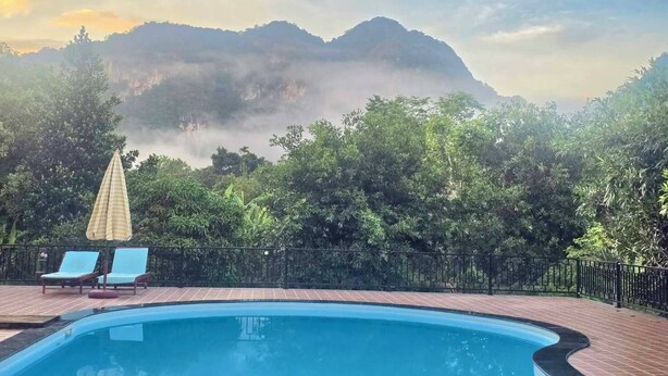 A swimming pool with lounge chairs and an umbrella, surrounded by trees and a mountain in the background. - Khao Sok Country Resort (Phanom)