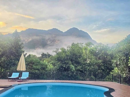 A swimming pool with lounge chairs and an umbrella, surrounded by trees and a mountain in the background.