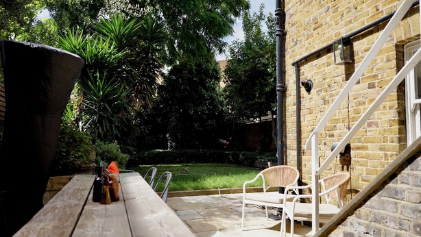A patio with a wooden table and chairs, a large planter, and a brick wall. - Cozy Victorian 1BDR Garden Flat in Greenwich (Greater London)