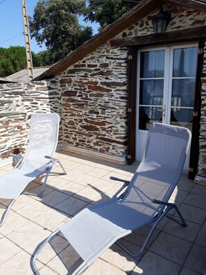 Ein Steinhaus mit einer gefliesten Terrasse, zwei weißen Liegestühlen und einer Glastür.