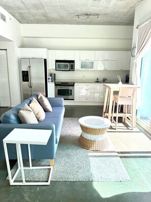 A modern kitchen with white cabinets, a refrigerator, microwave, and stove. There is a dining area with a table and chairs, and a blue sofa with cushions. - Water view apartment with free metro in the building (Miami)