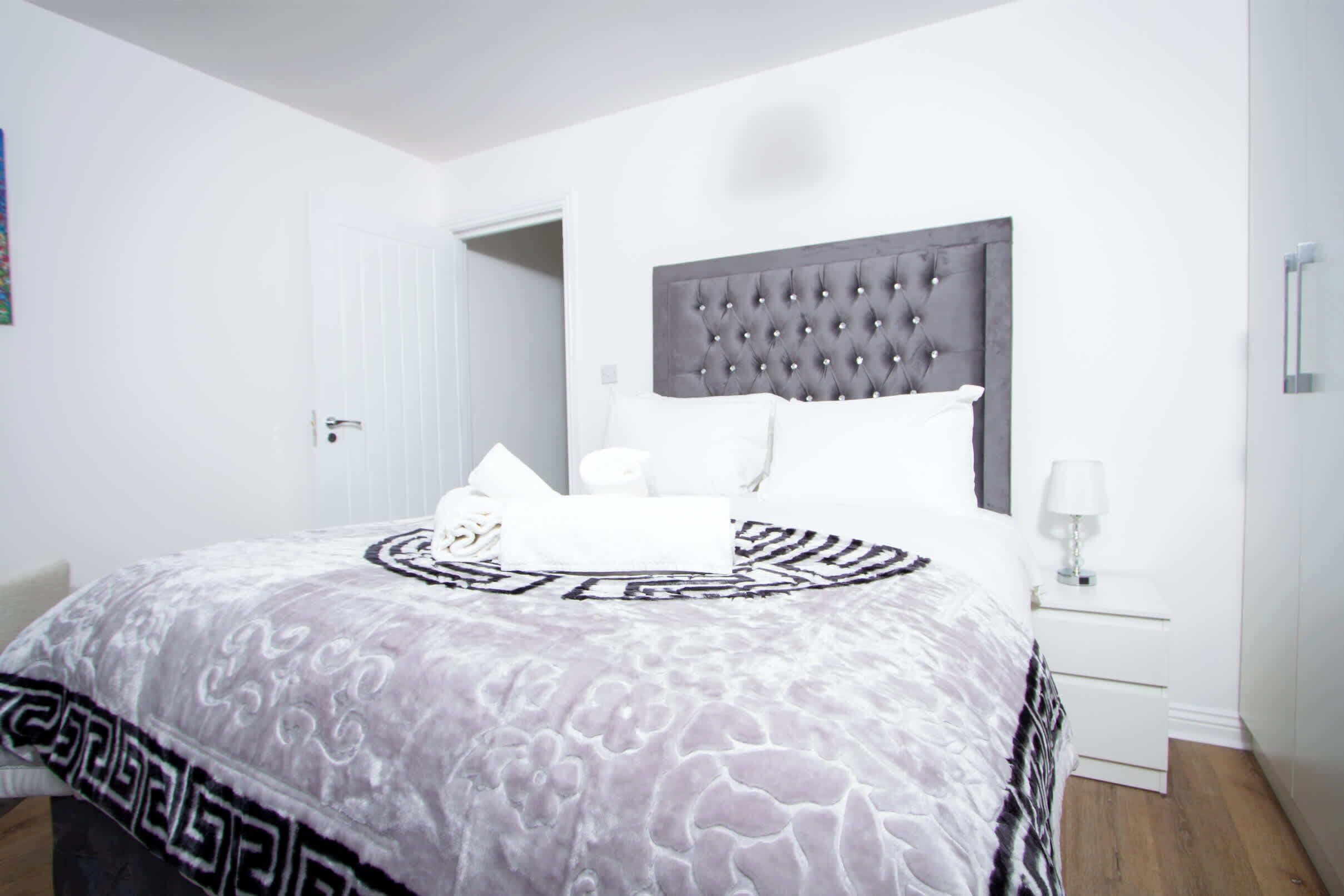 Un dormitorio con cabecera capitonada, ropa de cama blanca, una mesita de noche con lámpara y una puerta que da a otra habitación.