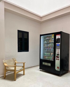 Ein Zimmer mit Getränkeautomaten, einem Fenster und einem Stuhl.