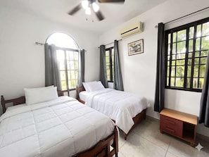 Une chambre avec deux lits, un ventilateur de plafond, un climatiseur, une fenêtre avec des rideaux et un petit banc en bois.