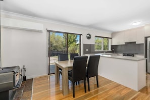 A modern kitchen with a central island, dining area with chairs, and large windows. - Rock On – Your Snowy Mountains Escape (Jindabyne)