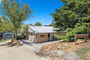 A small stone building with a metal roof, surrounded by trees and a gravel driveway. - Rock On – Your Snowy Mountains Escape (Jindabyne)
