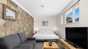 Habitació acollidora amb paret de pedra, llit amb roba de llit blanca, sofà gris, taula de centre de fusta i un televisor sobre un suport de fusta.