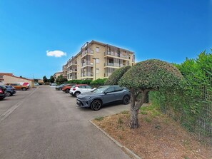 Ein Parkplatz mit mehreren Autos, ein Gebäude mit Balkonen und einer gepflegten Hecke.