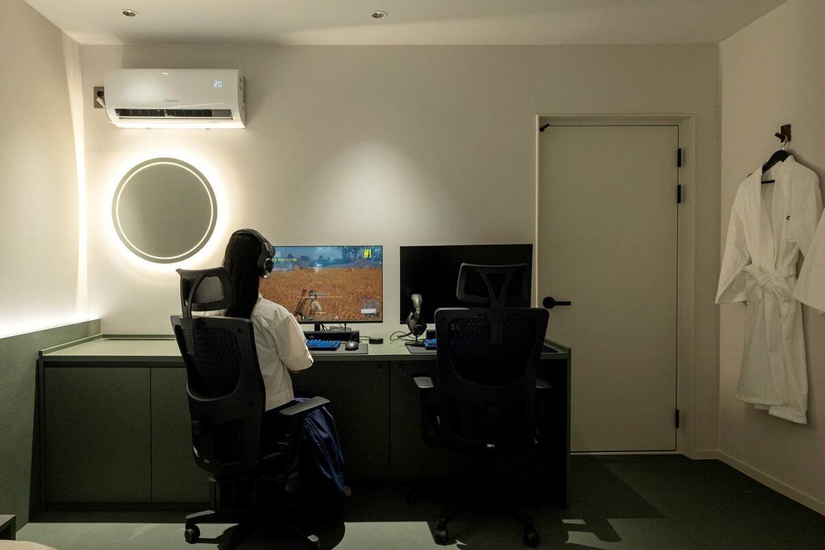 Une chambre d’hôtel équipée d’un bureau, d’un ordinateur et d’un grand écran. On y trouve un luminaire rond fixé au mur, un climatiseur installé au plafond et une porte avec un porte-serviettes.